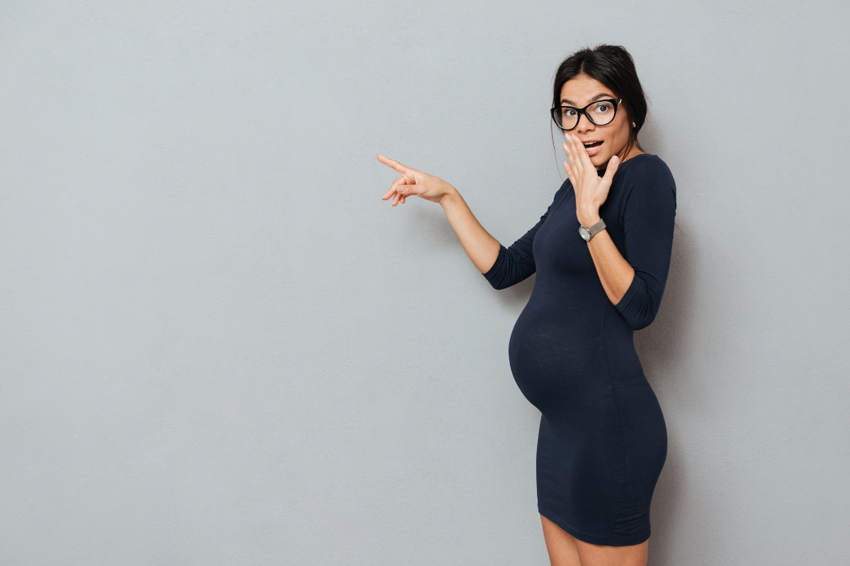 A pregnant woman wearing glasses and a dress looking shocked.